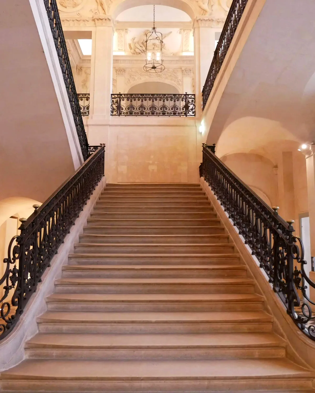 Un grand escalier de pierre claire à rampes de fer forgé, sous une lanterne